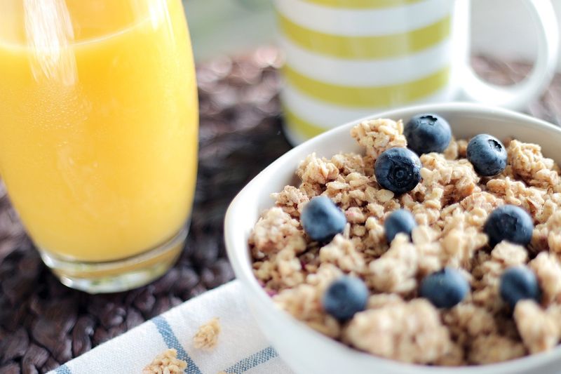 Photo d'un bol de muesli floconneux aux fruits ou fruits secs, sans sucre ajouté, accompagné d'un verre de jus d'orange.