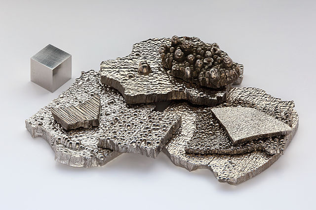 Fragments pures (à plus de 99,9 %) de cobalt, raffinés par électrolyse, à côté d’un cube d’1 cm³ de cobalt de haute pureté (99,8 %), pour comparaison. Attention : la comparaison est faussée du fait du polissage du cube seul.