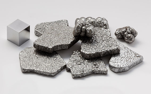 Fragments pures (à plus de 99,97 %) de fer, raffinés par électrolyse, à côté d’un cube d’1 cm³ de fer de haute pureté (99,9999 %), pour comparaison. Attention : la comparaison est faussée du fait du polissage du cube seul.